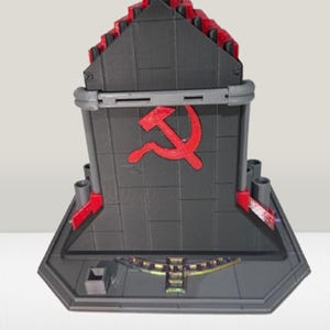Op de afbeelding: Een grijs en rood 3D-geprint model van een gebouw met een hamer en sikkel embleem. Het model heeft een geometrisch ontwerp met een rood dak en accenten. De basis is donkergrijs met een decoratief element.