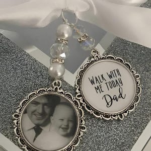 Op de afbeelding: Twee zilverkleurige bedels met een witte achtergrond. Een bedel heeft een foto van een vader en een kind. De andere bedel heeft de tekst "Loop vandaag met me mee Papa".