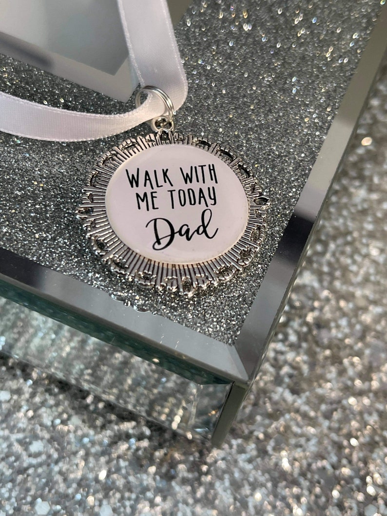 May include: A silver charm with a white background and black text that reads "Walk with me today Dad". The charm is attached to a white ribbon.