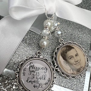 Puede incluir: Dos abalorios conmemorativos plateados con una cinta de satén blanca. Un abalorio presenta una foto en blanco y negro de un joven. El otro abalorio tiene el texto "Missing you as I walk down the aisle".