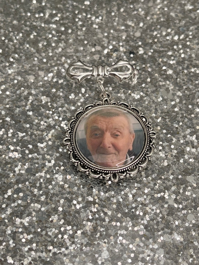 K&ouml;nnte beinhalten: Ein silberfarbener Foto-Anh&auml;nger mit einer Schleifen-f&ouml;rmigen Nadel an der Oberseite. Der Anh&auml;nger hat eine klare Glasfront und ein Foto eines Mannes im Inneren.