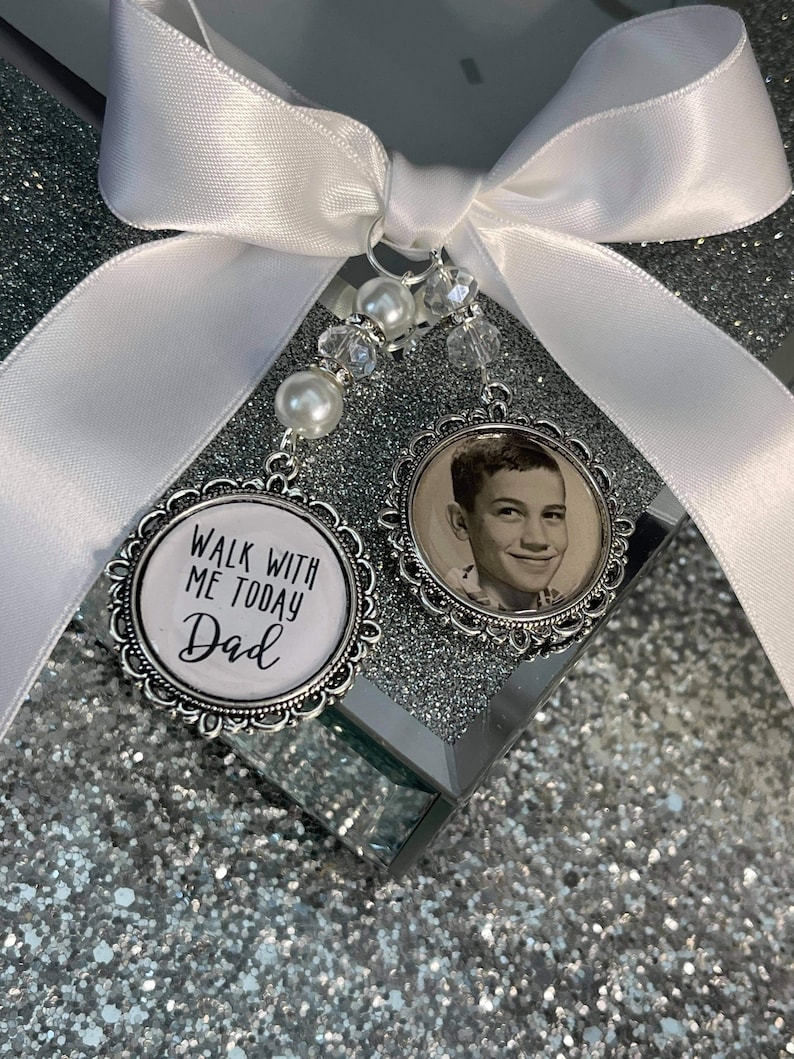 May include: A white satin ribbon tied into a bow with a silver charm attached. The charm has a black and white photo of a man and the text "Walk with me today Dad".