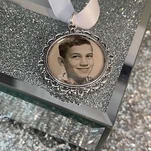 May include: A silver locket with a decorative border and a black and white photo of a young boy inside. The locket is hanging from a white ribbon.