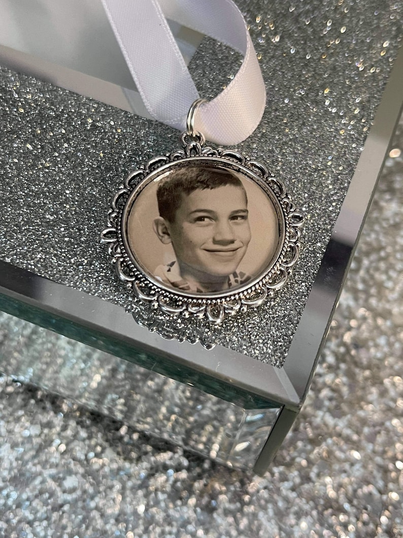 May include: A silver locket with a decorative border and a black and white photo of a young boy inside. The locket is hanging from a white ribbon.