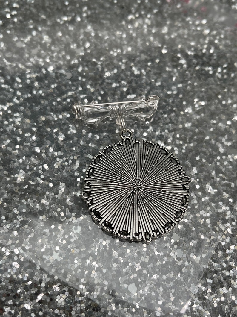K&ouml;nnte beinhalten: Silberfarbene Brosche mit einem runden Design mit einem Sonnenstrahlenmuster. Die Brosche hat eine Nadel auf der R&uuml;ckseite.