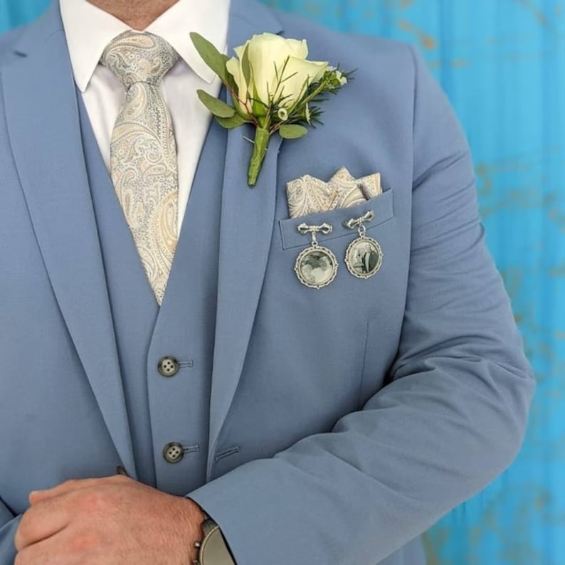 K&ouml;nnte beinhalten: Ein hellblauer Anzugsakko mit einem wei&szlig;en Blumenanstecker, einer gemusterten Krawatte und zwei silbernen Fotoanh&auml;ngern, die am Einstecktuch befestigt sind.