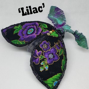 May include: A black fabric butterfly with purple flowers and green leaves. The butterfly has a glittery finish and the words "Lilac" are written above it.