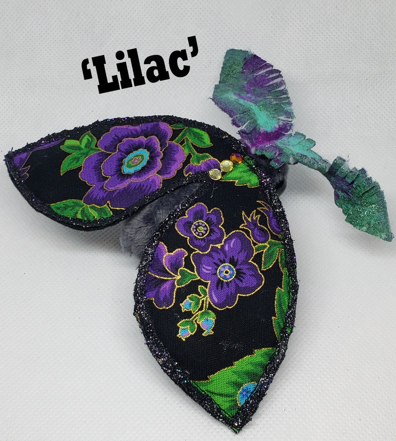May include: A black fabric butterfly with purple flowers and green leaves. The butterfly has a glittery finish and the words "Lilac" are written above it.
