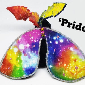 May include: A rainbow-colored fabric butterfly with a glittery trim and a black center. The butterfly has a yellow, orange, red, pink, blue, green, and purple color scheme. The butterfly has a glittery trim and a black center. The butterfly is decorated with rhinestones. The butterfly is labeled "Pride".