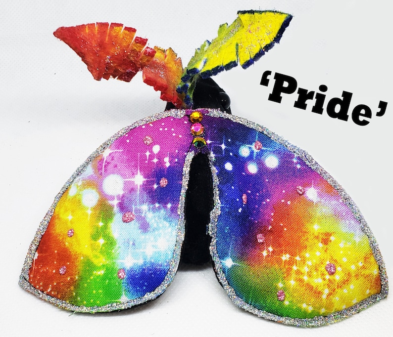 May include: A rainbow-colored fabric butterfly with a glittery trim and a black center. The butterfly has a yellow, orange, red, pink, blue, green, and purple color scheme. The butterfly has a glittery trim and a black center. The butterfly is decorated with rhinestones. The butterfly is labeled "Pride".