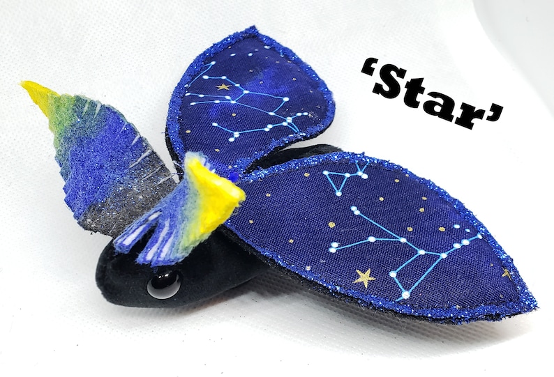 May include: A plush toy shaped like a bat with blue wings decorated with gold stars and constellations. The wings are outlined in gold glitter. The bat has a black body with yellow accents and a single black eye. The text 'Star' is written in black letters on a white background.