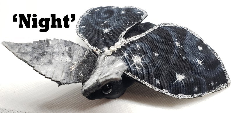 May include: A black fabric bat with silver glitter accents and a starry night sky design. The bat has a small black head with a single black eye. The text 'Night' is visible in the image.