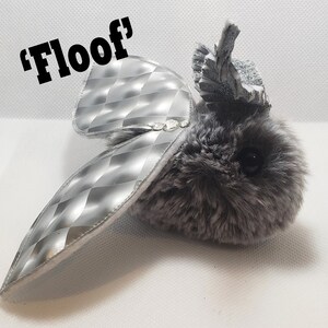 May include: A gray and silver plush toy with a silver, diamond-patterned wing. The toy has a crown-like headpiece and the text 'Floof' is written above it.
