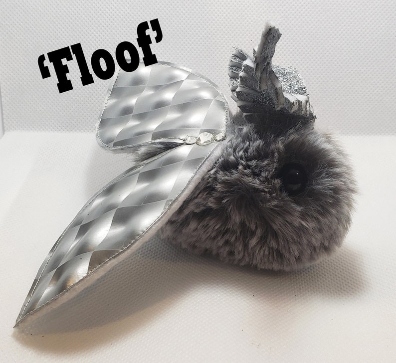 May include: A gray and silver plush toy with a silver, diamond-patterned wing. The toy has a crown-like headpiece and the text 'Floof' is written above it.