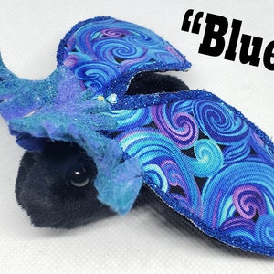 May include: A plush toy with a blue and purple swirl fabric design. The toy is shaped like a bat with large wings and a small body. The toy has a black face with large black eyes. The toy is labeled "Bluey".