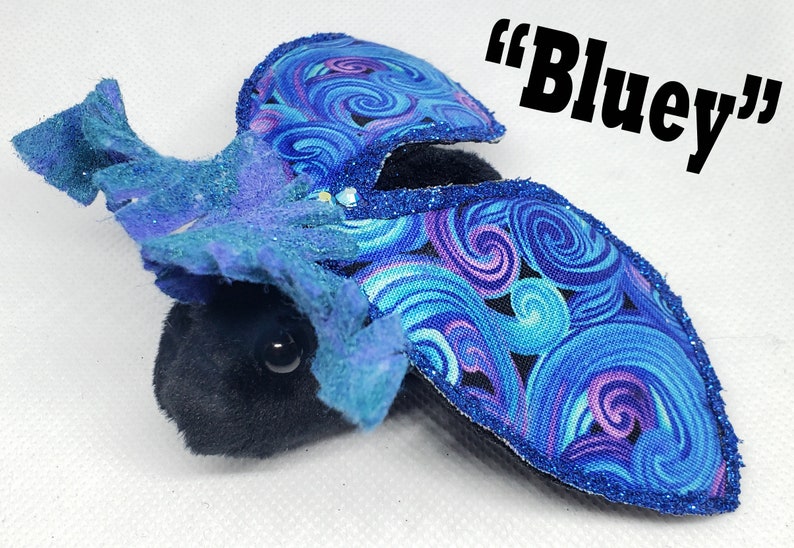 May include: A plush toy with a blue and purple swirl fabric design. The toy is shaped like a bat with large wings and a small body. The toy has a black face with large black eyes. The toy is labeled "Bluey".