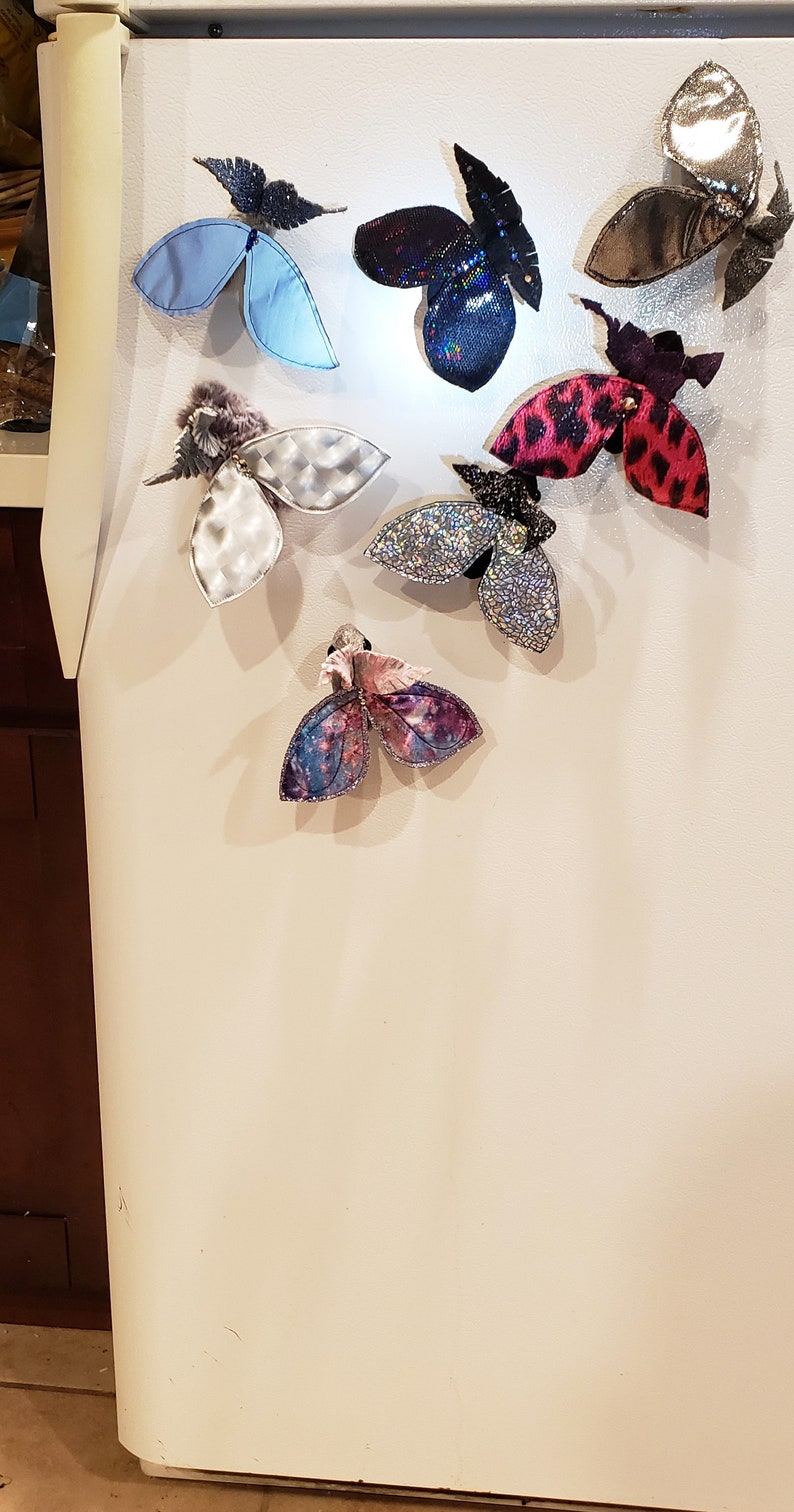 May include: A white refrigerator door with eight colorful butterfly magnets attached. The butterflies are made of fabric and have different patterns and textures.