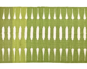 May include: A rectangular green rug with a white geometric pattern. The rug has a fringe on both ends. The pattern consists of vertical white shapes against a green background, with varying shades of green.
