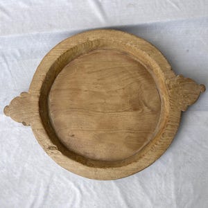 Op de afbeelding: Een ronde, lichtbruine houten schaal met een opstaande rand en decoratieve handvatten. De schaal heeft een natuurlijke houtnerf en een glad, onafgewerkt oppervlak. De handvatten hebben een gesneden, sierlijk ontwerp.