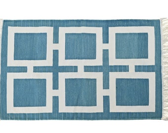 Puede incluir: Una alfombra rectangular con un diseño geométrico. La alfombra presenta un fondo azul turquesa con un patrón de cuadrados y líneas blancas. Los bordes tienen flecos blancos.