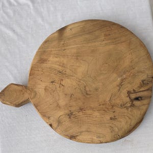 以下が含まれることがあります： ハンドル付きの丸い木製まな板。ボードは、木目がはっきりと見える、自然な仕上がりの明るい茶色の木材で作られています。ハンドルは長方形で、端はひし形です。