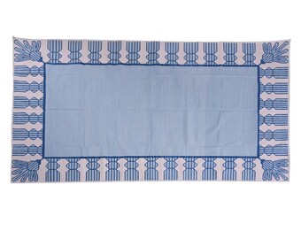 Puede incluir: Textil rectangular azul claro y blanco. El centro es azul claro liso, enmarcado por un borde de diseños geométricos azules y blancos. Los bordes presentan motivos florales estilizados.
