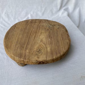 Op de afbeelding: Een ronde, lichtbruine houten serveerplank met een natuurlijk houtnerfpatroon. De plank heeft een licht verhoogde rand en drie kleine voetjes. Het hout heeft een warme, natuurlijke tint, perfect voor het presenteren van voedsel of decoratie.