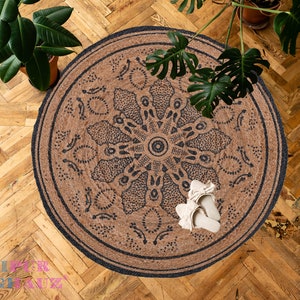 May include: A round jute rug with a black and white mandala design. The rug is on a wooden floor with a plant in a pot in the upper left corner and another plant in a pot in the upper right corner.