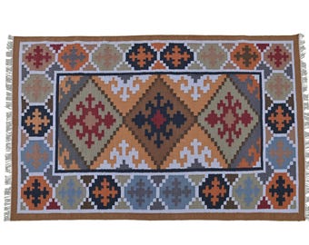 May include: Rectangular woven rug with a geometric pattern in shades of brown, orange, red, and blue. The rug has a brown border and fringe on the short ends. The central design features diamond and cross shapes.