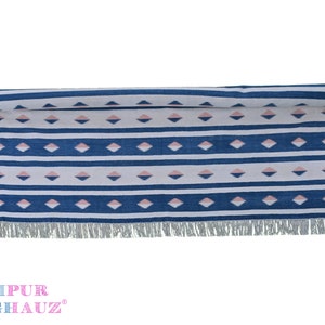 May include: A blue and white striped rug with pink diamond patterns and fringe on the ends. The rug has the text "JAIPUR RUGHAUZ" on the bottom.