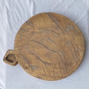 以下が含まれることがあります： 丸い木製のまな板、取っ手付き。明るい茶色の木材でできており、木目と質感がよく見えます。取っ手は長方形で穴が開いています。白い表面に置かれています。