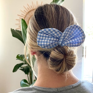 Könnte beinhalten: Ein blaues und weißes Gingham-Haarband in Form einer Schleife. Die Schleife ist zu einem Knoten gebunden und wird im Haar einer Person mit blonden Haaren getragen.
