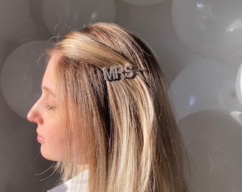 Könnte beinhalten: Ein silberner Haarschmuck mit dem Wort "MRS" in Blockbuchstaben. Der Haarschmuck ist im blonden Haar einer Frau befestigt. Weiße Ballons sind im Hintergrund.
