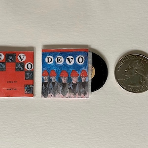 May include: A red and blue vinyl record with a picture of a band called DEVO on the cover. The record is next to a quarter for size comparison.