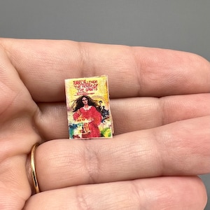 May include: A miniature paperback book with a yellow cover and a colorful illustration of a woman in a red dress holding a birdcage. The title of the book is "The House of the Spirits" by Isabel Allende.