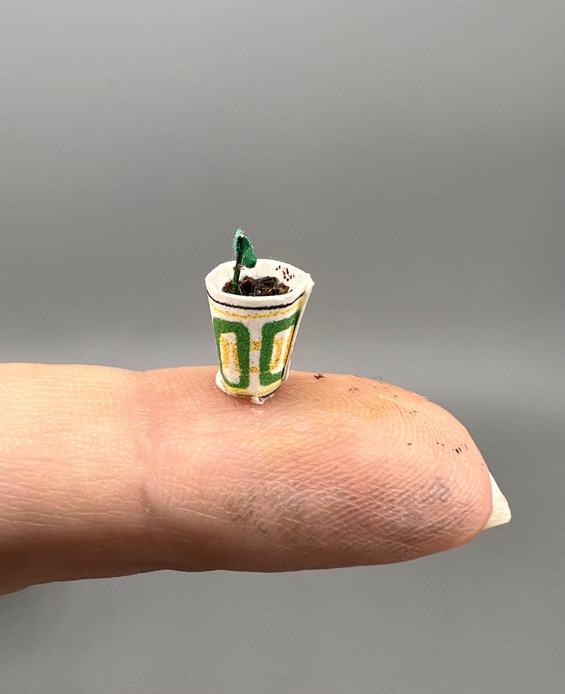 May include: A miniature potted plant with a green stem and leaves, planted in brown soil. The pot is made of paper and has a green and yellow geometric pattern.