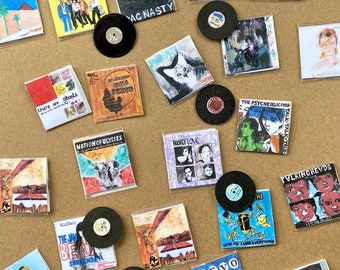 Puede incluir: Una colección de portadas de álbumes en miniatura y discos de vinilo. Las portadas presentan varios nombres de bandas y arte de álbumes, incluyendo "The B-52's", "Jawbreaker" y "Devo". Los discos son negros con etiquetas blancas. Los artículos están dispuestos sobre una superficie marrón claro.