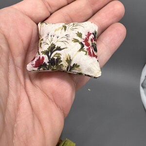 Puede incluir: Una peque&ntilde;a almohada cuadrada con un patr&oacute;n floral en tonos rojos, verdes y blancos. La almohada est&aacute; hecha de una tela suave y blanca y tiene un brillo sutil.