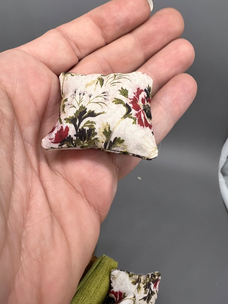 Puede incluir: Una peque&ntilde;a almohada cuadrada con un patr&oacute;n floral en tonos rojos, verdes y blancos. La almohada est&aacute; hecha de una tela suave y blanca y tiene un brillo sutil.