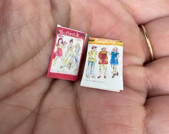May include: Two vintage sewing pattern envelopes, one red with "Butterick" printed on it and the other white with "Simplicity" printed on it. Both envelopes feature illustrations of women's clothing.