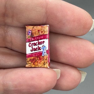 May include: Miniature Cracker Jack box with a red and white label. The box features the text "The Original Cracker Jack" and a cartoon image of a sailor. The box has a brown and gold speckled pattern.