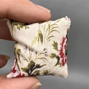Puede incluir: Una peque&ntilde;a almohada blanca con un patr&oacute;n floral en tonos rojos, verdes y marrones. La almohada est&aacute; hecha de tela y tiene un borde ligeramente deshilachado.