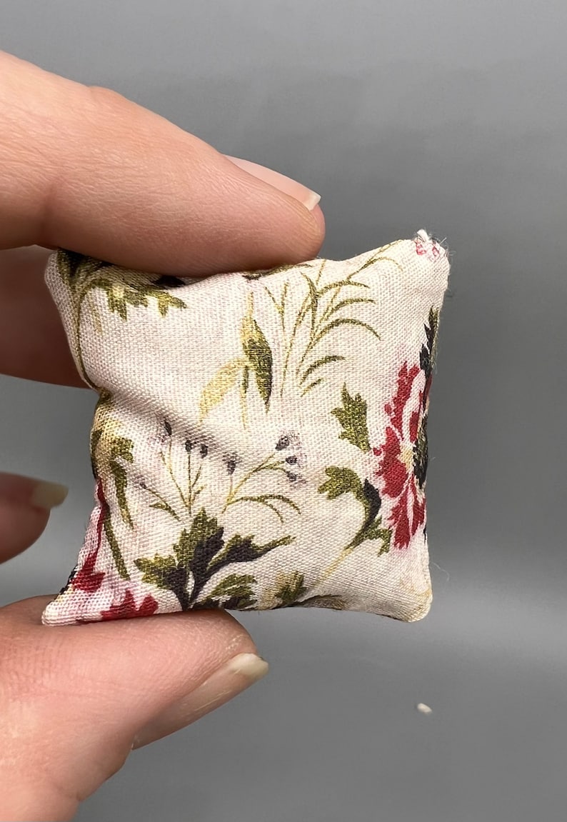 Puede incluir: Una peque&ntilde;a almohada blanca con un patr&oacute;n floral en tonos rojos, verdes y marrones. La almohada est&aacute; hecha de tela y tiene un borde ligeramente deshilachado.