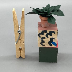 May include: A wooden clothespin with a metal spring, a small potted plant with green leaves, and a stack of three painted wooden blocks. The top block is pink with black and blue abstract designs, the middle block is beige with black abstract designs, and the bottom block is dark green.