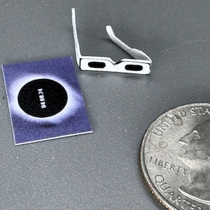 Puede incluir: Un par de gafas de eclipse blancas con lentes negros. Las gafas están sobre una superficie gris junto a una pequeña tarjeta con una imagen de un eclipse solar y la fecha "040824" impresa en ella. También se puede ver una moneda de 25 centavos de dólar estadounidense en la imagen.