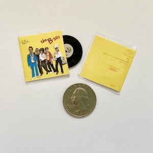 May include: A yellow vinyl record album cover with the band "The B-52's" printed on the front. The album cover features a photo of the band members in colorful clothing. The album cover is next to a black vinyl record and a yellow paper sleeve.