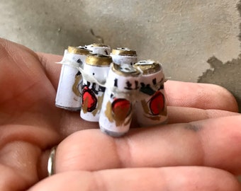 May include: A close-up of six miniature cans of beer, each with a white label and a red apple design. The cans are arranged in a circle and are held in a person's hand.
