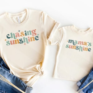 May include: Two cream-colored t-shirts, one adult and one child size, with the phrases "Chasing Sunshine" and "Mama's Sunshine" in colorful retro fonts. The shirts are paired with blue jeans, suggesting a casual, family-oriented theme.