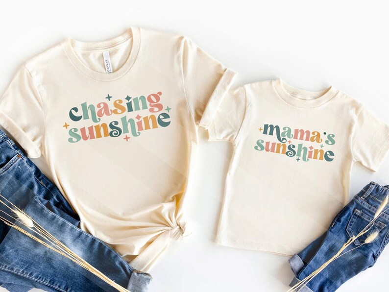 May include: Two cream-colored t-shirts, one adult and one child size, with the phrases "Chasing Sunshine" and "Mama's Sunshine" in colorful retro fonts. The shirts are paired with blue jeans, suggesting a casual, family-oriented theme.