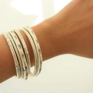 May include: A set of six silver bangles with etched designs. The bracelets are stacked on a wrist, showcasing their intricate patterns and reflective surface. The jewelry is displayed against a soft, neutral background.